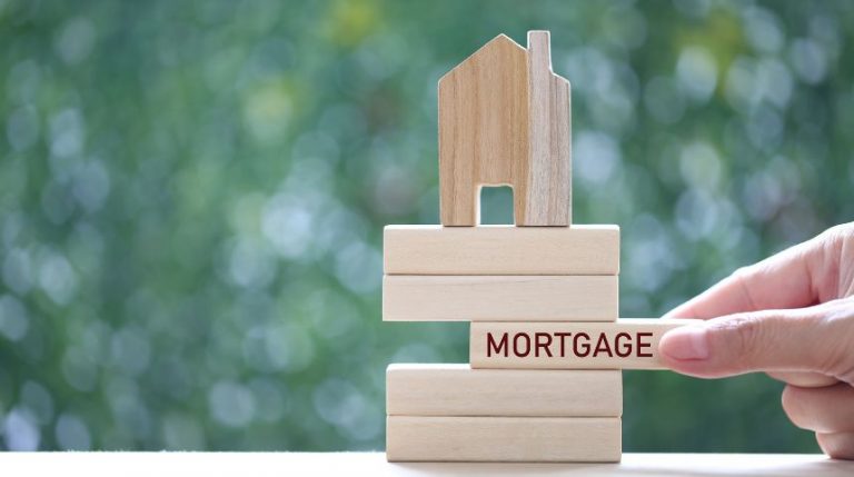 A stack of wooden blocks with a small wooden house on top. A hand is pulling out a piece in the middle with the word "mortgage" written on it.