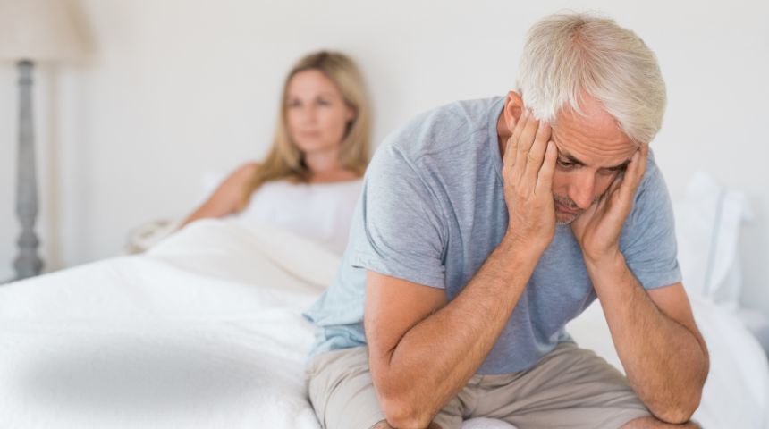 An older couple in bed, with the man sitting holding his head, looking frustrated.