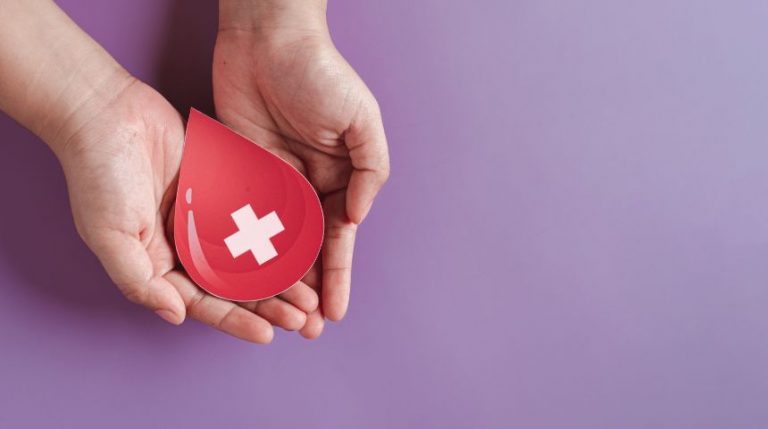 Two hands holding a paper cut out of a red drop of blood with a white cross on it, against a purple background.