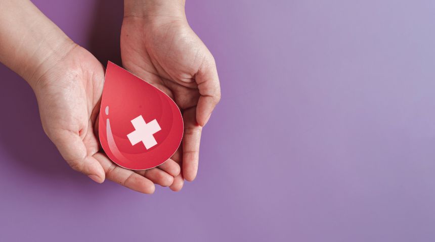 Two hands holding a paper cut out of a red drop of blood with a white cross on it, against a purple background.